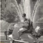 1960 lady reading paper on Lambretta scooter in frond of fountain